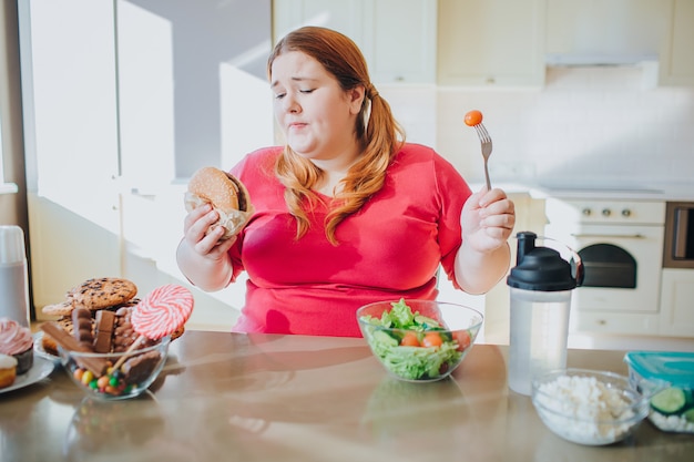 Mujer joven gorda en la cocina que se sienta y que come la comida basura.