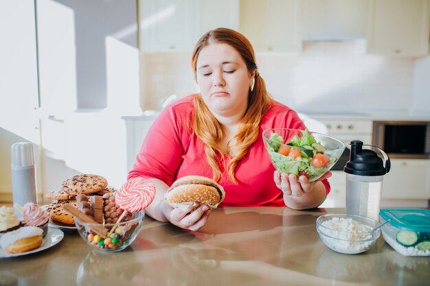 Mujer joven gorda en la cocina que se sienta y que come la comida. Estilo de vida saludable.