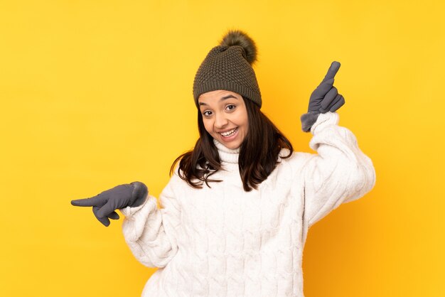 Mujer joven con gorro de invierno sobre fondo amarillo aislado dedo acusador hacia los laterales y feliz