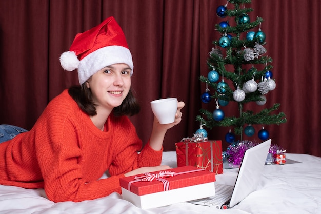 Una mujer joven con un gorro de Papá Noel y un suéter rojo sostiene una taza de café o té, descansa con su computadora portátil, acostada en la cama, rodeada de regalos de Navidad. Compras navideñas online