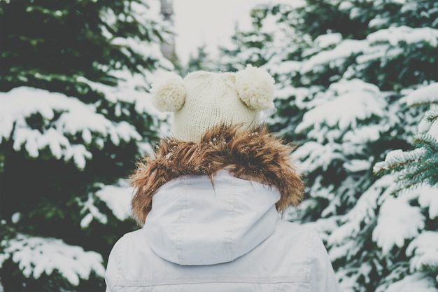 Mujer joven con gorro de punto va entre abetos en un bosque.