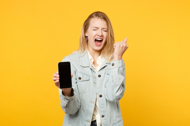 Mujer joven gritando con ropa informal de denim apretando el puño, sosteniendo un teléfono móvil con una pantalla vacía en blanco aislada en un fondo amarillo anaranjado en el estudio. Concepto de estilo de vida de las personas. Simulacros de espacio de copia.