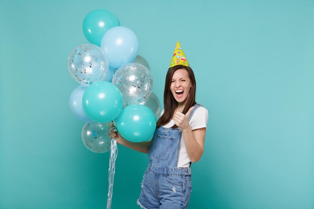 Mujer joven gritando con ropa de mezclilla, sombrero de cumpleaños mostrando el pulgar hacia arriba celebrando, sosteniendo coloridos globos de aire aislados en un fondo azul turquesa. Fiesta de cumpleaños, concepto de emociones de la gente.