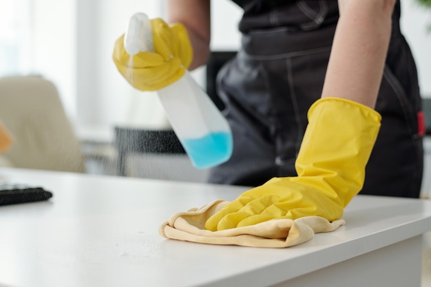 Mujer joven en guantes de goma amarillos protectores limpiando el polvo en el escritorio