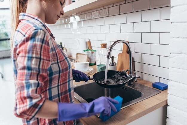 Mujer joven con guantes de goma lavando una sartén en la cocina.