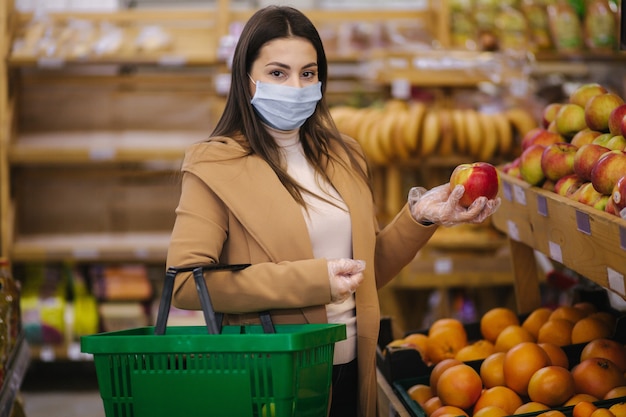 Mujer joven con guantes protectores y mascarilla sostiene hermosa manzana fresca en la mano. Hermosa joven con canasta de alimentos eligiendo alimentos por stand con frutas. Compras durante la cuarentena. COVID-19