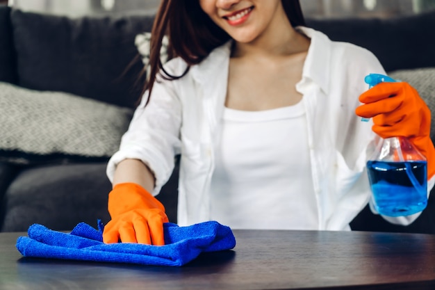Mujer joven en guantes protectores con un spray y un trapo mientras limpia la casa en la sala de estar en casa.