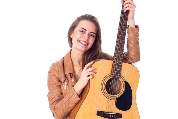 Mujer joven guapa con chaqueta marrón sosteniendo una guitarra con fondo blanco en el estudio