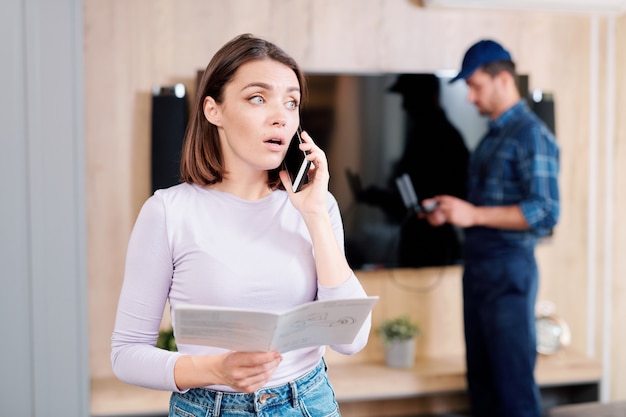 Mujer joven con guía de instrucciones de uso que consulta el servicio de reparación del hogar en el teléfono