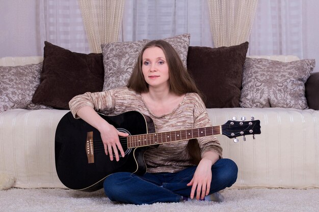 Mujer joven con guitarra acústica negra sobre una alfombra en la habitación