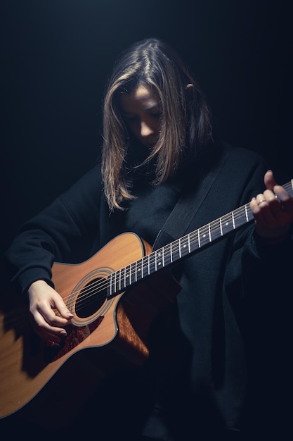 Una mujer joven con una guitarra acústica en la oscuridad bajo un rayo de luz.