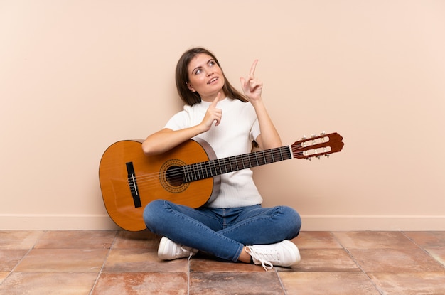 Mujer joven con guitarra sentada en el suelo señalando con el dedo índice una gran idea