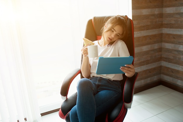 Mujer joven en la habitación. Siéntate en una silla y trabaja de forma remota. Hablar por teléfono y mirar la tableta en las manos al mismo tiempo. Empresaria fuera de la oficina en casa.