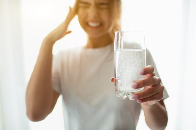 Mujer joven en la habitación. Sufre de fuerte dolor de cabeza o dolor. Sostenga el vaso con una mesa efervescente. Resaca o dolor de cabeza.