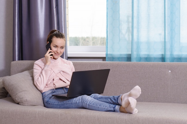 Mujer joven hablando por teléfono trabajando simultáneamente en una computadora portátil sentada en el sofá de la habitación