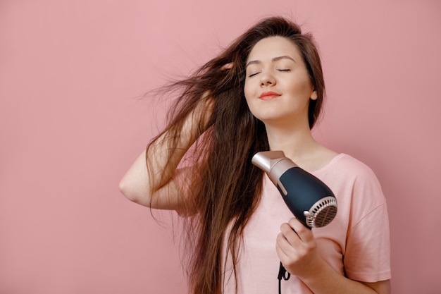 Mujer joven hace volumen de cabello con secador de pelo en mano sobre fondo rosa