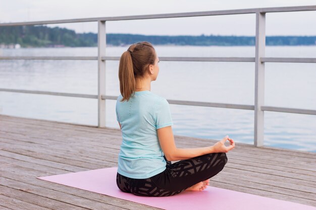 Mujer joven hace yoga en una alfombra rosa en un muelle junto al mar