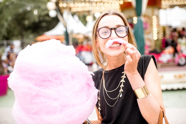 Mujer joven haciendo bigote con algodón de azúcar rosa de pie al aire libre en el parque de atracciones