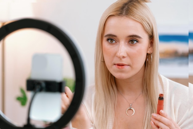 Mujer joven haciendo contenido de video tutorial de belleza y cosméticos para redes sociales Blogger de belleza usando cámara y anillo de luz mientras muestra cómo aplicar lápiz labial líquido a la audiencia o seguidor Blithe