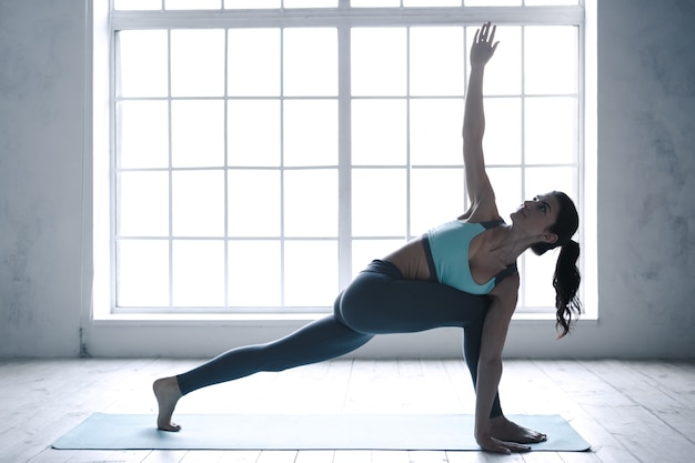 Mujer joven haciendo ejercicio de asana de yoga en el interior cerca de la ventana