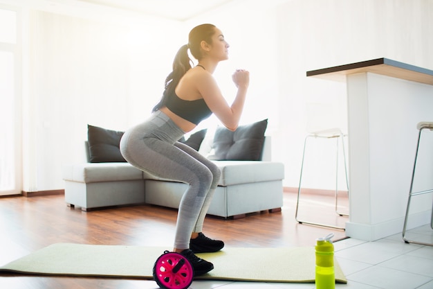 Mujer joven haciendo ejercicio deportivo en la habitación durante la cuarentena. Haciendo ejercicios de sentadillas sobre colchoneta de yoga en la habitación. Entrenamiento concentrado.