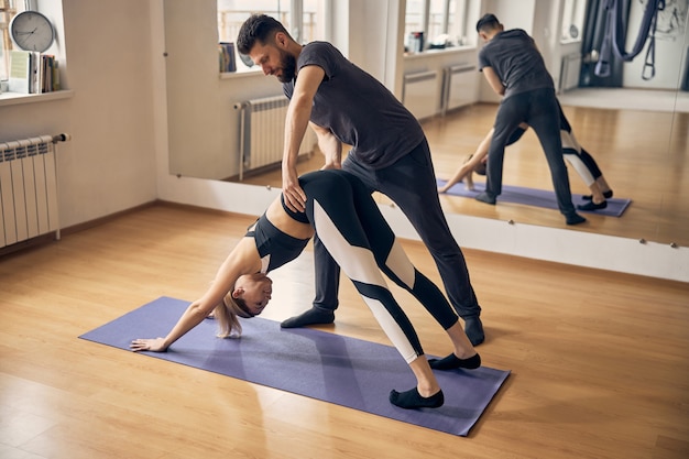 Mujer joven haciendo ejercicio en una estera de yoga especial mientras un instructor experimentado la ayuda con la pose