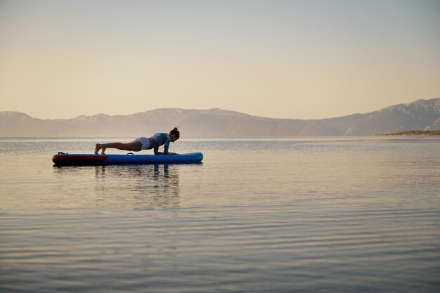 Mujer joven haciendo ejercicio de tablones en una tabla de remo flotando en el agua tranquila de la mañana.