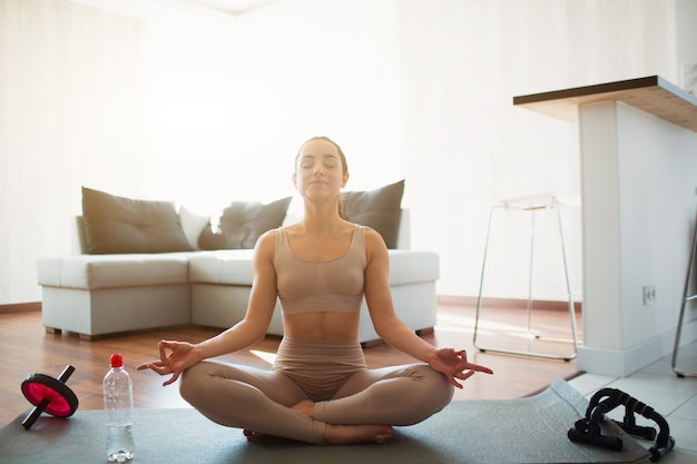 Mujer joven haciendo ejercicios de yoga en la habitación durante la cuarentena. Siéntese en la colchoneta en posición de loto con las piernas cruzadas. Meditando solo en la habitación. Botella de agua y equipamiento deportivo para el hogar además.