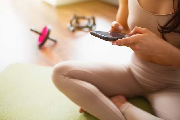 Mujer joven haciendo ejercicios de yoga en la habitación durante la cuarentena. Vista de corte de niña con smartphone. La mujer se sienta en la estera durante el descanso.