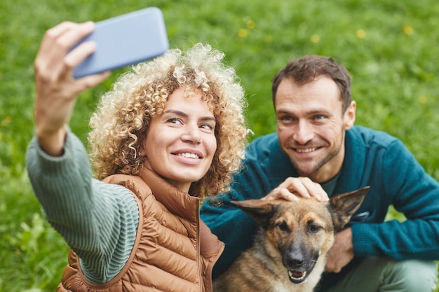 Mujer joven haciendo retrato selfie en su teléfono móvil con perro y su novio al aire libre