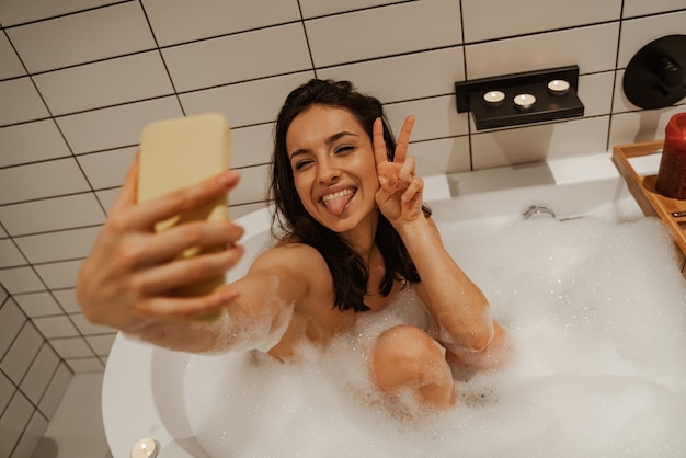 Mujer joven haciendo selfie mientras se relaja en el hermoso baño lleno de espuma decorado con velas. Trato saludable y disfrute