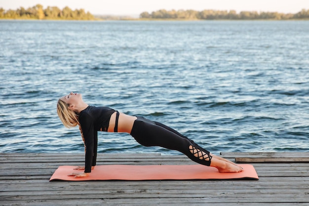 Foto una mujer joven está haciendo un tablón inverso en una colchoneta de yoga junto al río.