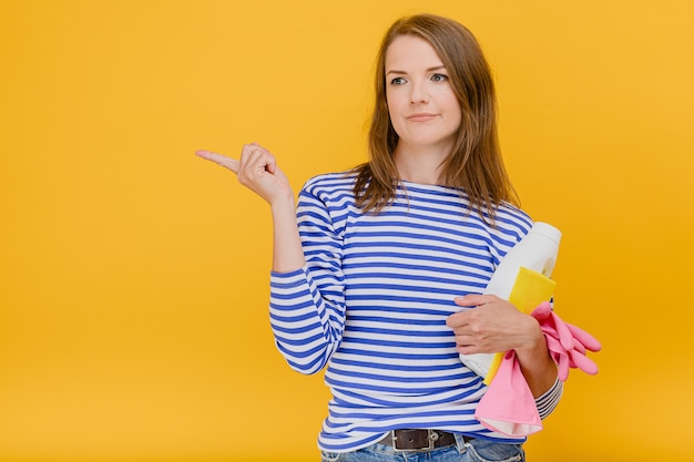 Mujer joven haciendo tareas domésticas sosteniendo detergente con guantes protectores de goma esponja se preocupa de ropa sanitaria casual suéter de rayas azules