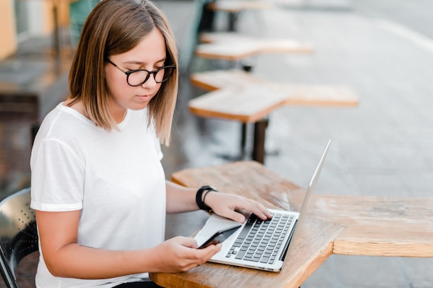 Mujer joven haciendo trabajo independiente desde el teléfono y la computadora portátil en la cafetería al aire libre