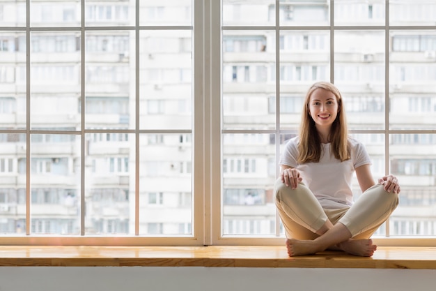 Mujer joven haciendo yoga delante de la ventana. Mujer feliz practicando yoga mientras está sentado en el alféizar de la ventana en posición de loto asana