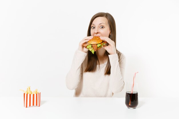 Mujer joven hambrienta come con avidez hamburguesa, se sienta a la mesa con papas fritas, cola en botella aislada sobre fondo blanco. Nutrición adecuada o comida rápida clásica estadounidense. Área de publicidad con espacio de copia.