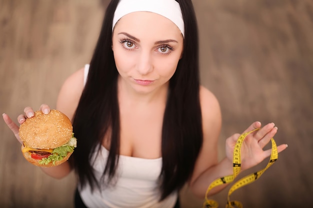 Una mujer joven con una hamburguesa y una cinta métrica. Una niña se encuentra en una madera. La vista desde la cima. El de una alimentación saludable.