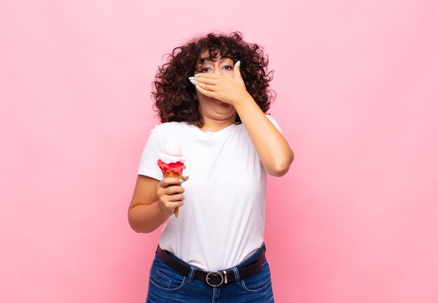 Mujer joven con un helado cubriendo la boca con las manos con una expresión de asombro y sorpresa, guardando un secreto o diciendo uy