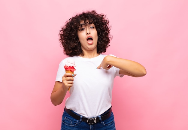 Mujer joven con un helado mirando consternado y sorprendido con la boca abierta, apuntando a sí mismo