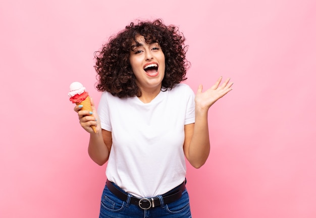 Mujer joven con un helado que se siente feliz, emocionada, sorprendida o conmocionada, sonriendo y asombrada por algo increíble