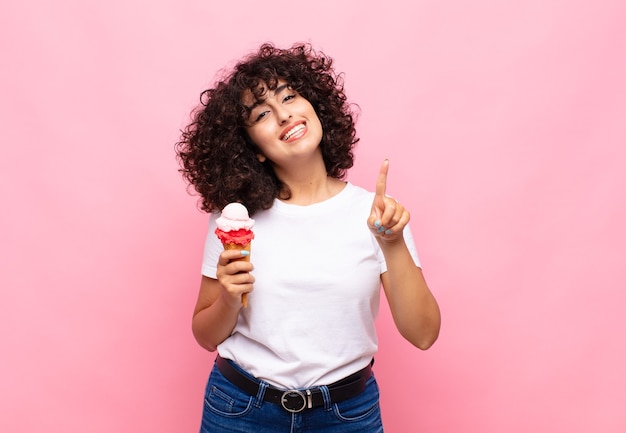 Mujer joven con un helado sonriendo y mirando amistosamente, mostrando el número uno o primero con la mano hacia adelante, contando hacia atrás