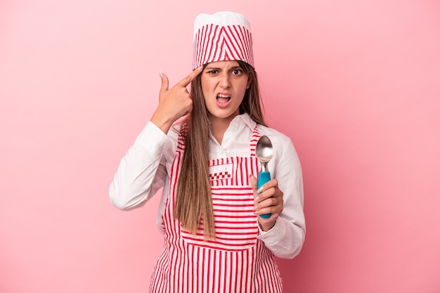 Mujer joven heladora sosteniendo una cuchara aislada sobre fondo rosa mostrando un gesto de decepción con el dedo índice.