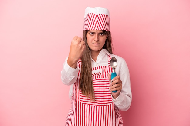 Mujer joven heladora sosteniendo una cuchara aislada sobre fondo rosa mostrando el puño a la cámara, expresión facial agresiva.