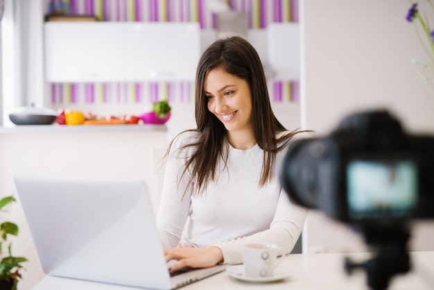 La mujer joven hermosa alegre está usando su computadora portátil y está tomando café delante de una cámara.