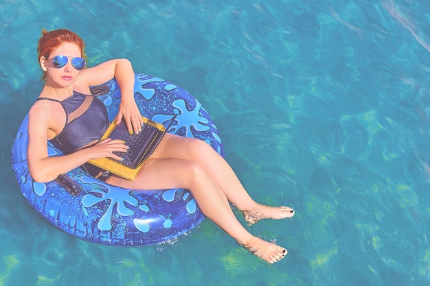 Mujer joven hermosa con un anillo inflable y una computadora portátil que se relajan en el mar.