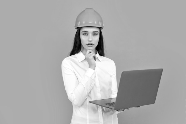 Mujer joven hermosa arquitecta con casco de sombrero duro sostenga la computadora portátil sobre fondo gris
