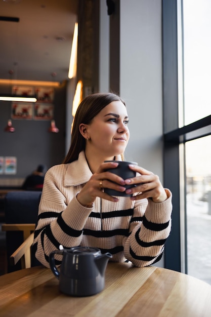 Una mujer joven y hermosa bebe café y mira por la ventana mientras está sentada en un café en una mesa junto a la ventana La mujer joven disfruta de la mañana Taza de café vigorizante por la mañana