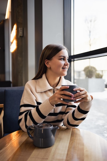 Una mujer joven y hermosa bebe café y mira por la ventana mientras está sentada en un café en una mesa junto a la ventana La mujer joven está disfrutando de la mañana Taza de café vigorizante por la mañana