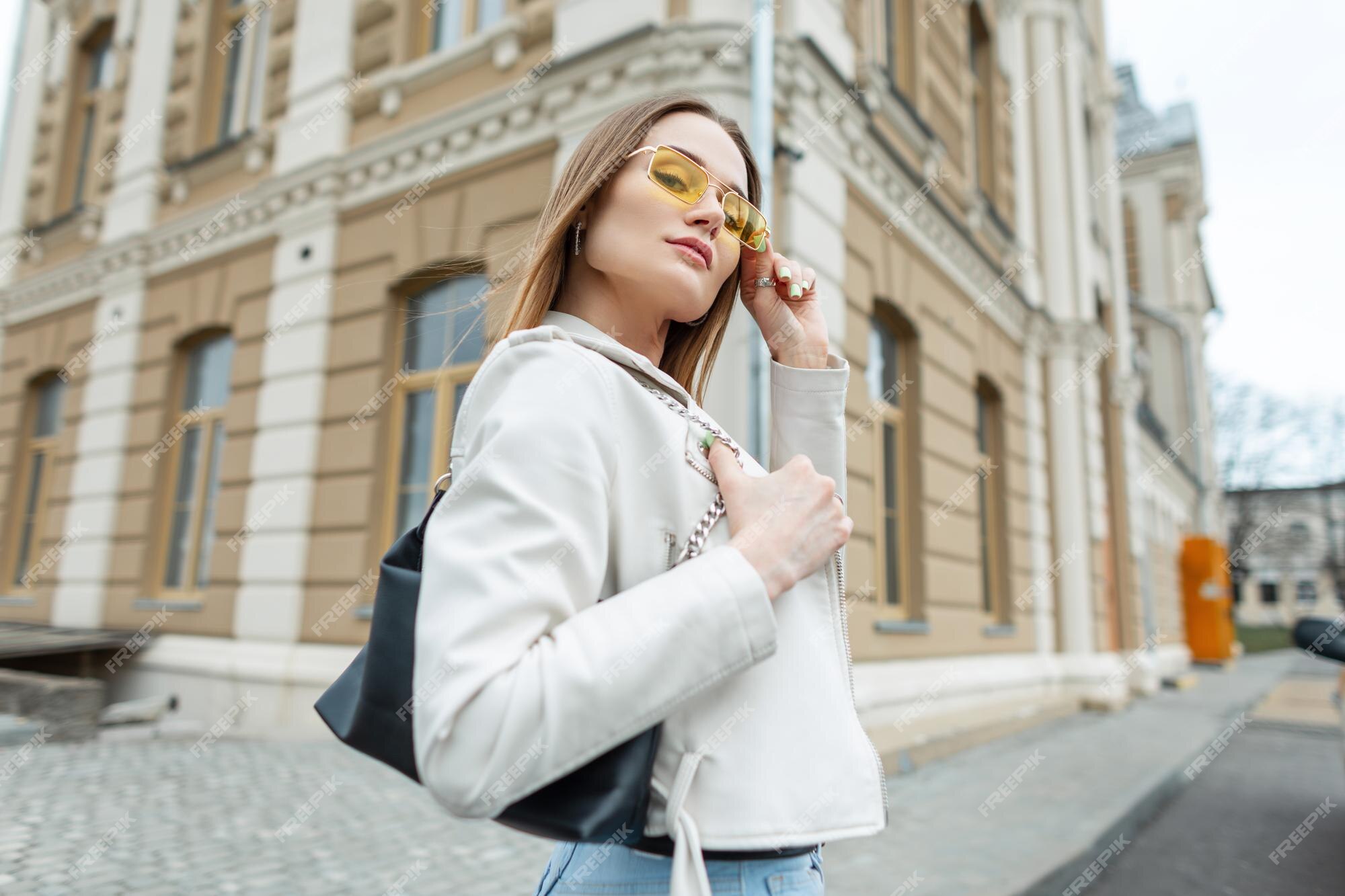 Mujer joven hermosa y elegante con gafas de sol vintage amarillas en ropa de moda con jeans de chaqueta de y paseos de cartera en la ciudad y mira a la