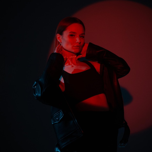 Mujer joven hermosa y elegante con ropa negra de moda posando sobre un fondo oscuro en un estudio con luz roja creativa